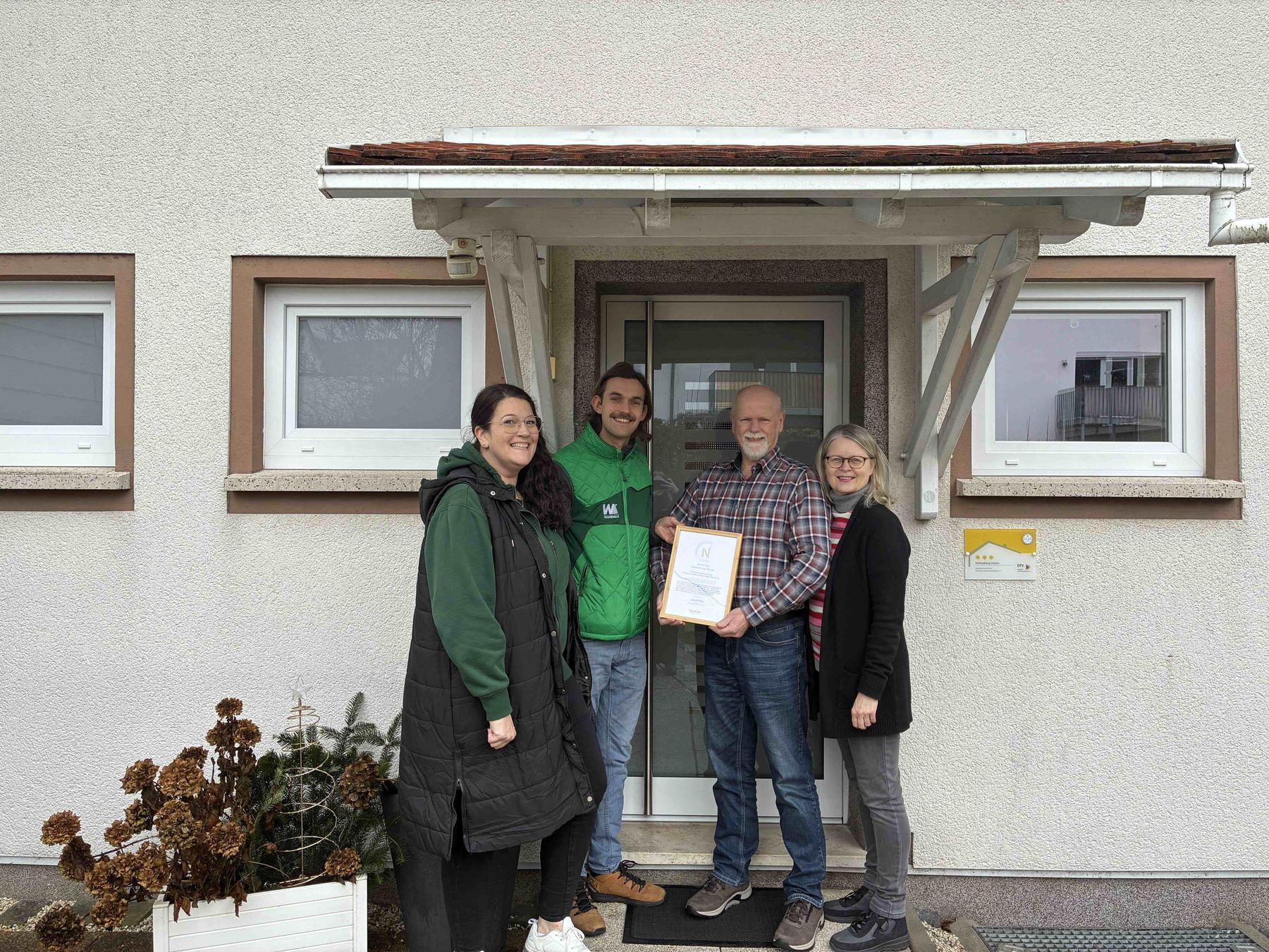 Four people stand smiling in front of a building entrance. One person in the center holds a framed certificate. There are two windows, a white planter with dried flowers, and a canopy above the door.
